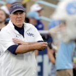 Bill Belichick watches his team warm up on the field before a game, wearing a navy "Tar Heels" ball cap and a white UNC quarter zip.