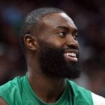 Jaylen Brown smiles, looking forward on the court, wearing a green jersey.