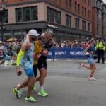 Israel Rivera (L) and Henco Visser (R) carry Jonathan Adams along Boylston Street at the 2026 Boston Marathon.