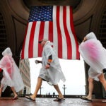 Pedestrians in rain ponchos walk by a huge American flag.