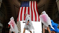 Pedestrians in rain ponchos walk by a huge American flag.