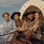 "A man and three women in pioneer-era clothing ride a horse-drawn covered wagon outdoors, under a blue sky with clouds, suggesting a journey or travel in a historical rural setting."
