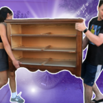 A woman and a man carrying a brown empty dresser with no drawers. The woman is wearing a black sports bra and black shorts, and the man is wearing a black t-shirt and black and white shorts. The background is faded to purple, and there's a white glow behind the dresser.