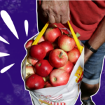 A man's hand is holding a big white bag of red apples. He's wearing a red shirt and jeans. The background is of the grass in the orchard, faded to purple, and there are white accent marks around the bag of apples.
