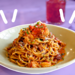 Viet-Cajun crawfish noodles from Le Madeline in a white bowl, outlined in white. The table has a red iced drink behind it, which is all faded into a light purple background. There are three white accent marks on the left and right sides of the noodles.