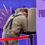 A man in a checkered collared shirt, jeans, and suspenders, voting in a polling station, hunched over a table with a divider. He and his station are outlined in white, and there is a red divider rope next to him. The background is of more polling areas in the station, and it's faded to purple. There are white accent lines on the left of his back.