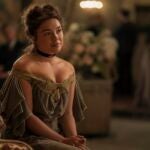 "A woman in a vintage off-shoulder dress sits on a patterned sofa in a softly-lit, elegant room with blurred people and floral arrangements in the background, suggesting a formal or historic gathering."