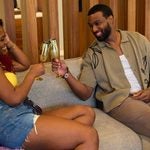 "Two people sit on a couch in a cozy living room, smiling and toasting with gold glasses. They relax among purple pillows, suggesting a casual, friendly atmosphere."