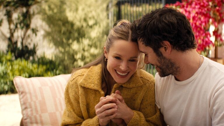 "A smiling woman in a yellow sweater and a man in a white shirt sit closely together outside on a patio, surrounded by lush greenery and vibrant pink flowers, appearing happy and affectionate."